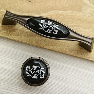 May include: Two cabinet knobs with a black and white floral design. The larger knob is a pull handle with a black ceramic insert and a bronze metal base. The smaller knob is round with a black ceramic insert and a bronze metal base.
