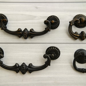 May include: Four antique-style cabinet pulls in black metal with ornate designs. Two are arched pulls with a decorative center piece, and two are ring pulls with a round base.