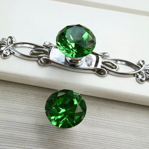 Pode incluir: Dois botões de cristal verde para armário com base prateada adornada. Os botões são facetados e têm forma de diamante.