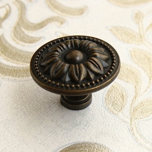 May include: A bronze-colored cabinet knob with a floral design. The knob has a round base with a raised, detailed flower design in the center. The knob has a small, round stem.