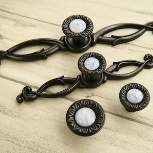 Może przedstawiać: Trzy ozdobne czarne metalowe gałki do szafek z białymi wkładkami z porcelany. Gałki mają ozdobny wzór w postaci zwijającego się ornamentu.