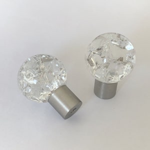 Può includere: Due pomelli per armadi in vetro trasparente con un design testurizzato a ghiaccio screpolato. I pomelli hanno una base in metallo argentato.