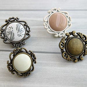 Puede incluir: Cuatro pomos decorativos para armario con marcos de metal ornamentados. Dos pomos tienen inserciones de cerámica blanca, uno tiene una inserción de cerámica beige y uno tiene una inserción de latón.