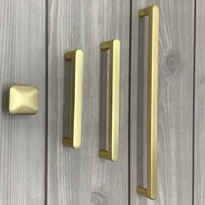 Könnte beinhalten: Vier goldfarbene Schrankgriffe in verschiedenen Größen und Stilen. Die Griffe sind auf einer hellgrauen Holzplatte montiert.