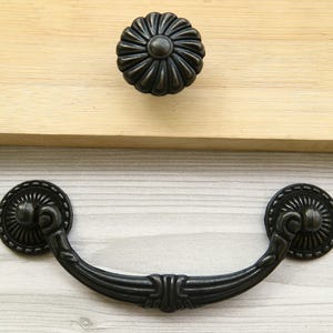 May include: Black metal drawer pull with a decorative floral design. The pull has a curved shape and two round ends.