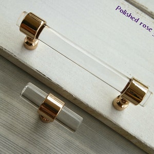 May include: Clear acrylic cabinet pulls with polished rose gold accents. The image shows a long pull and a smaller knob. The text "Polished rose gold" is visible in the upper right corner. These pulls are suitable for drawers and cabinets.