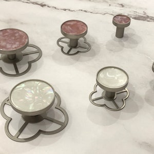 Puede incluir: Cinco pomos decorativos para armario con acabado plateado y base en forma de flor. Dos pomos tienen un diseño marmoleado rosa y blanco, y tres pomos tienen un diseño marmoleado blanco e iridiscente.
