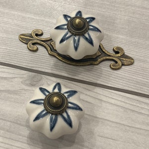 May include: Two white ceramic cabinet knobs with a blue floral design and a brass center. The knobs are shown on a light wood surface.