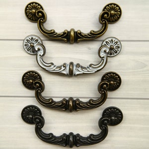 May include: Three ornate cabinet pulls in different finishes: antique brass, silver, and black. Each pull features a decorative floral design and a curved handle.
