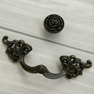 May include: Two antique bronze cabinet pulls with a floral design. One pull is a round knob with a rose design. The other pull is a curved handle with a rose design at the center.