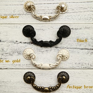 May include: Four ornate drawer pulls in different finishes: antique silver, black, white with gold accents, and antique bronze. Each pull has a decorative, curved design.