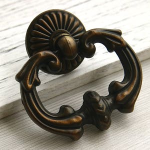 May include: Antique brass cabinet pull with a decorative, ornate design. The pull has a ring-shaped handle and a round, detailed base.