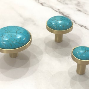 May include: Three turquoise and brass cabinet knobs with a round, flat base and a round, raised top. The knobs are different sizes.