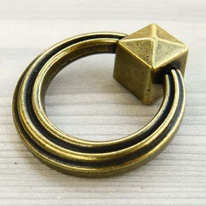 May include: A gold-toned ring pull cabinet handle with a square, faceted knob. The ring is double-layered and has a smooth, polished finish.