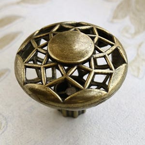 Puede incluir: Un pomo de armario de bronce con un diseño geométrico. El pomo es circular con un centro elevado y un patrón decorativo calado alrededor del borde.