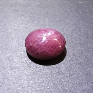 May include: A polished, oval-shaped gemstone with a deep pinkish-red hue. The surface reflects light, showcasing subtle internal patterns. The gem is smooth and appears to be a natural stone, possibly a ruby or similar precious stone.