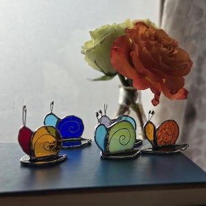 May include: Five colorful stained glass snails on a stack of books. The snails are made of glass and have different colors, including red, orange, yellow, blue, and green.