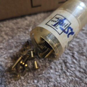 May include: A clear plastic container with a gold-colored rim, partially tipped over, spilling out numerous small, brass-colored tacks. The container has a label with blue handwritten text and a logo. The tacks are scattered on a textured, light brown surface.