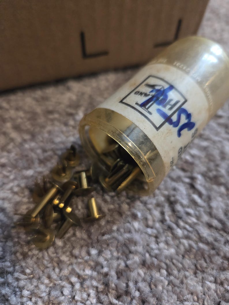 May include: A clear plastic container with a gold-colored rim, partially tipped over, spilling out numerous small, brass-colored tacks. The container has a label with blue handwritten text and a logo. The tacks are scattered on a textured, light brown surface.