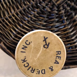 May include: A vintage, cylindrical container with a beige lid. The lid features the text "DERICK & REA INC." in a dark blue, circular font, along with the Rx symbol. The container is set against a woven basket background.