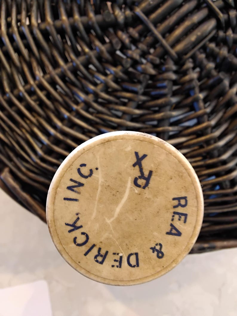 May include: A vintage, cylindrical container with a beige lid. The lid features the text "DERICK & REA INC." in a dark blue, circular font, along with the Rx symbol. The container is set against a woven basket background.