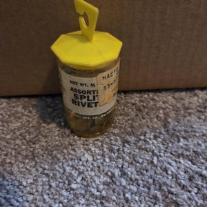 Puede incluir: Un pequeño recipiente de plástico transparente con tapa y asa amarillas, lleno de remaches partidos surtidos. La etiqueta dice "Assorted Split Rivet". El recipiente está sobre una alfombra gris.