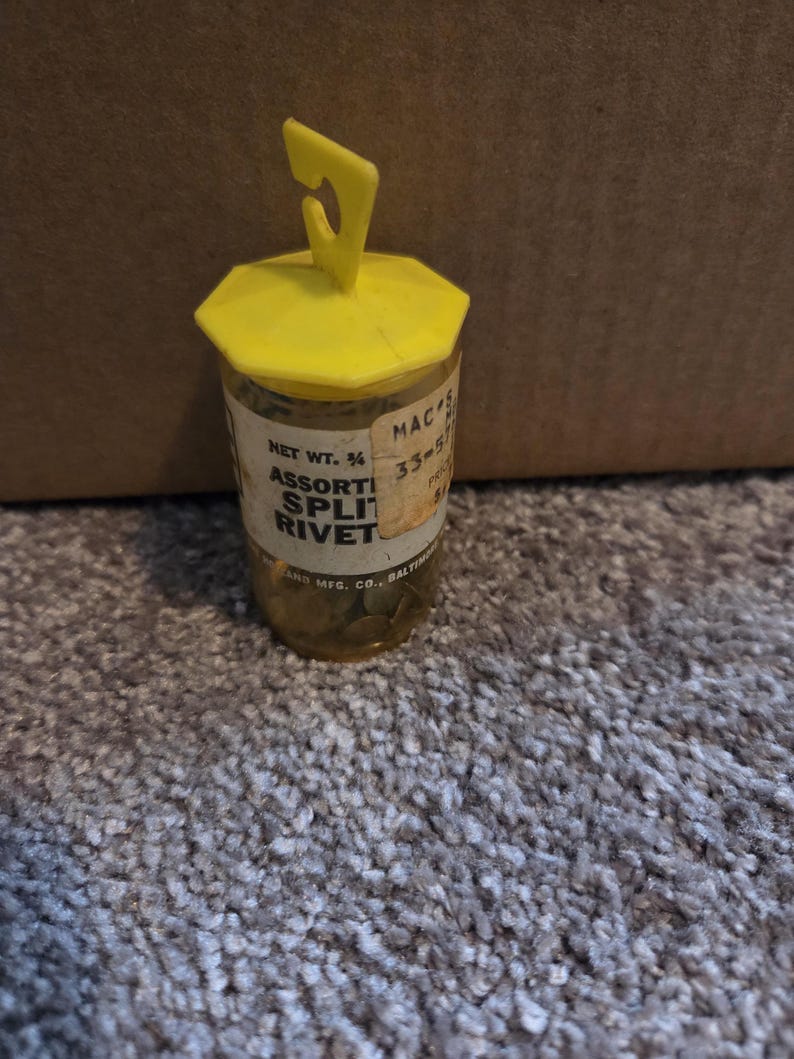May include: A small, clear plastic container with a yellow lid and handle, filled with assorted split rivets. The label reads "Assorted Split Rivet" and "Net Wt. 3/4". The container is on a gray carpet.