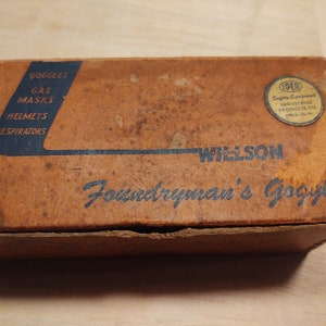 May include: A brown cardboard box with the text "Wilson Foundryman's Goggle" printed on the front. The box also has the text "Goggles, Gas Masks, Helmets, Respirators" printed on the side. The box has a label with the text "SPED Safety Equipment Industrial Products Co. Phila. Pa."