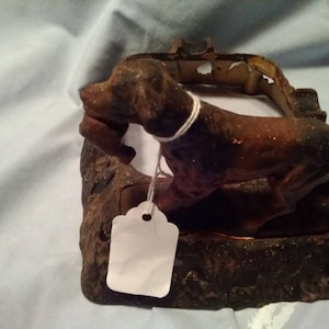 May include: A cast iron dog figurine with a brown and black finish. The dog is holding a smaller figurine in its mouth and is standing on a base with a rough texture.