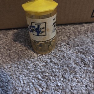 May include: A clear plastic container with a yellow lid and hanger, filled with small, round, gold-colored objects. The container has a label with text and a logo. The container is sitting on a gray carpet.