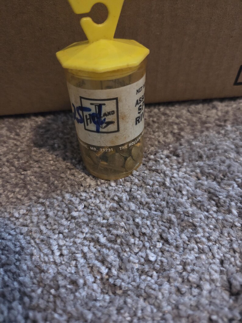May include: A clear plastic container with a yellow lid and hanger, filled with small, round, gold-colored objects. The container has a label with text and a logo. The container is sitting on a gray carpet.