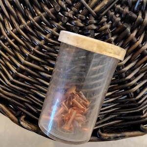 May include: A clear plastic container with a beige lid, filled with small copper-colored rivets. The container rests on a dark brown woven wicker surface, creating a contrast in textures and colors. The rivets appear to be new.