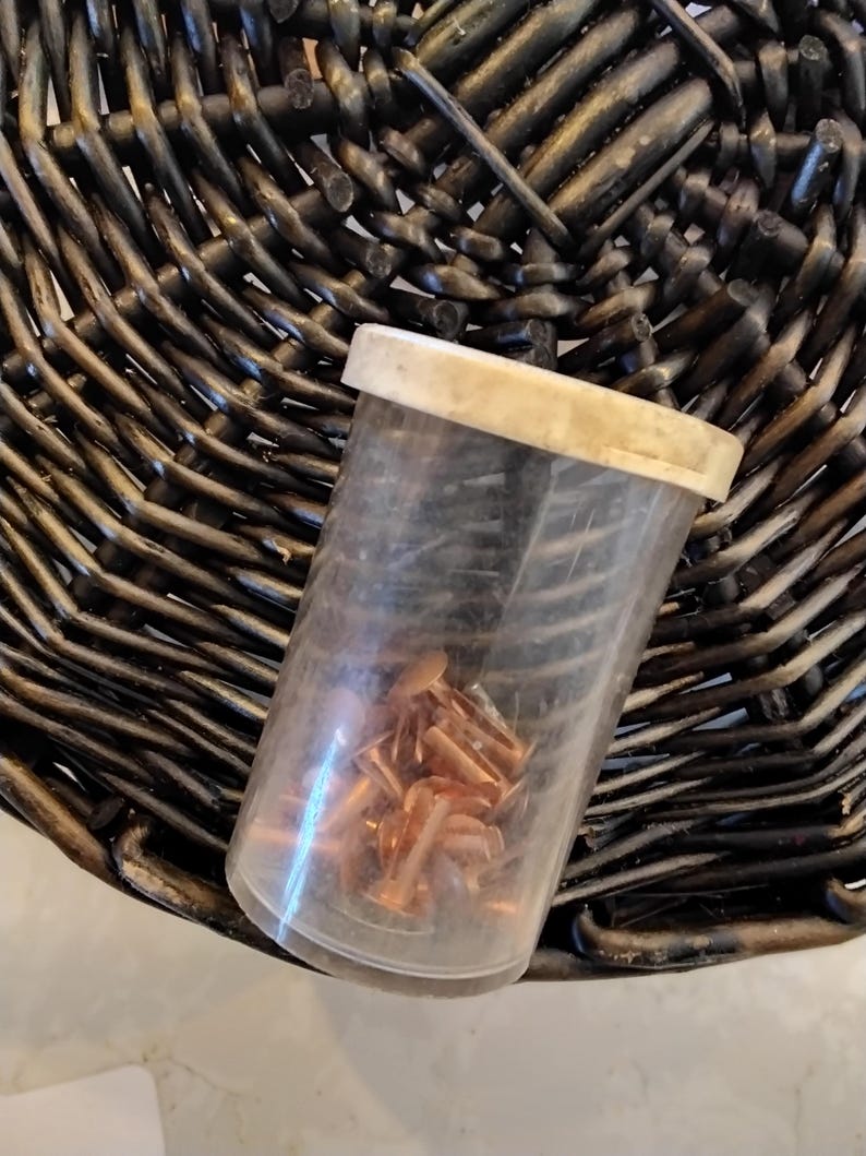 May include: A clear plastic container with a beige lid, filled with small copper-colored rivets. The container rests on a dark brown woven wicker surface, creating a contrast in textures and colors. The rivets appear to be new.