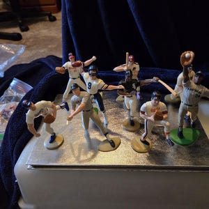 Puede incluir: Colección de figuras en miniatura de jugadores de béisbol en varias poses de acción. Las figuras visten diferentes uniformes de equipo, con detalles como guantes, bates y pelotas de béisbol. Las figuras están sobre bases redondas.
