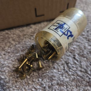 May include: A clear plastic container of brass-colored rivets, with some rivets spilled out onto a gray carpet. The container has a label with the text "25". A cardboard box is in the background.