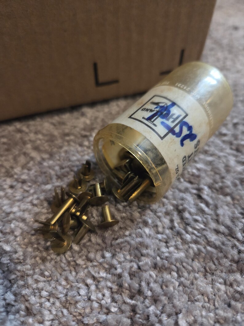 May include: A clear plastic container of brass-colored rivets, with some rivets spilled out onto a gray carpet. The container has a label with the text "25". A cardboard box is in the background.