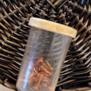 May include: A clear plastic container with a white lid, filled with small copper-colored metal objects. The container sits on a dark brown woven basket.