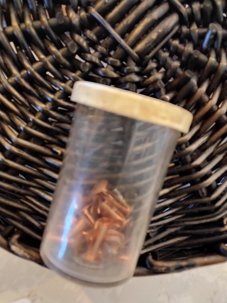 May include: A clear plastic container with a white lid, filled with small copper-colored metal objects. The container sits on a dark brown woven basket.