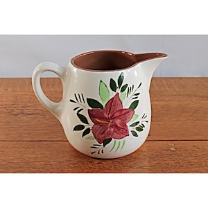 May include: A cream-colored ceramic pitcher featuring a hand-painted design of a large dark red flower with green leaves. The pitcher has a curved handle on the left side, a wide pouring spout on the right, and a smooth, glossy finish throughout.
