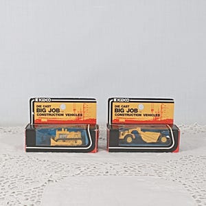 May include: Two die-cast toy construction vehicles in their original boxes. The boxes are black with red trim and feature the text "KIDCO DIE CAST BIG JOB... CONSTRUCTION VEHICLES". One box contains a yellow bulldozer, the other a yellow dump truck.