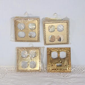 May include: Four square gold-toned metal wall plates for electrical outlets. Three plates are sealed in clear plastic packaging with hanging tabs, while one plate is unwrapped, featuring an ornate embossed border and two circular openings.