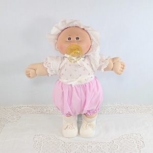 May include: A vintage Cabbage Patch Kid doll, featuring a soft, fabric body and a vinyl head. The doll wears a white bonnet, a floral-print top, pink bloomers, white socks, and white sneakers. It has a pacifier in its mouth.