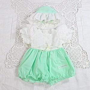 May include: A mint green and white baby outfit set, including a romper, bonnet, and top. The romper has a ruffled hem and a small label. The top has short sleeves with lace trim and a small floral pattern. The bonnet has a ruffled brim.