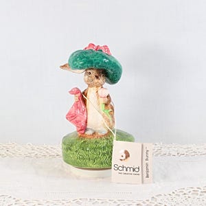 May include: A ceramic figurine of Benjamin Bunny, wearing a green hat with a pink bow and holding a pink bag. The figurine is on a green base, with a Schmid tag attached. The figurine is a collectible item.