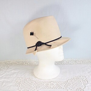 May include: A cream-colored cloche hat with a black band and decorative accents. The hat features a small black square and a pin on the side, with a knotted detail and triangular shapes on the brim. The hat is displayed on a white mannequin head.