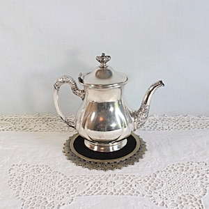 May include: A polished silver teapot featuring a curved handle, a decorative spout with intricate detailing, and a matching lid with a small finial. The teapot rests on a circular, dark-colored base with a scalloped, metallic edge.