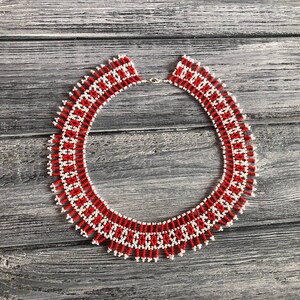 May include: A red and white beaded necklace with a geometric pattern. The necklace is made of small, round beads and has a fringe-like design at the bottom.