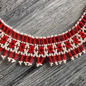 May include: A red and white beaded necklace with a fringe design. The necklace is made of small, round beads that are strung together in a geometric pattern.