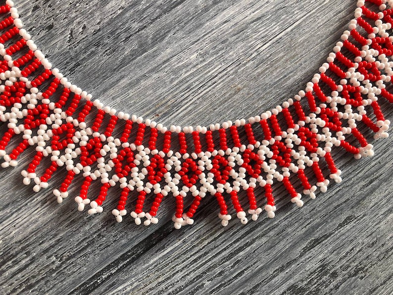 May include: A red and white beaded necklace with a fringe design. The necklace is made of small, round beads that are strung together in a geometric pattern.