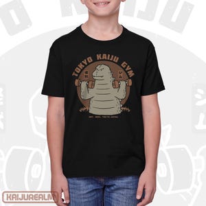 May include: Black t-shirt featuring a cartoon Godzilla lifting weights with the text "TOKYO KAIJU GYM" in a circular design. The shirt also includes the text "EST. 1954, TOKYO JAPAN".